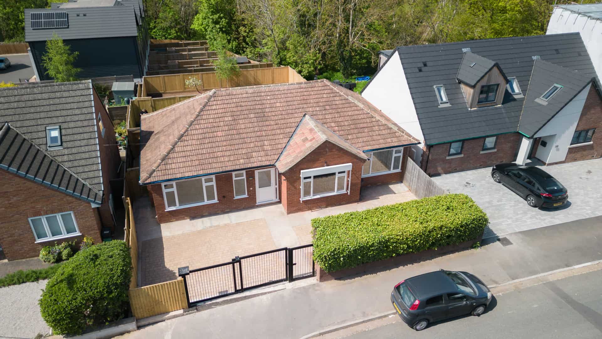 Aerial view of a charming single-story brick house with a tiled roof, nestled between two modern homes. A driveway and small front yard adorn this residence, while parked cars line the street. Perfect blend of classic and contemporary in this picturesque neighborhood of Purpose Homes.