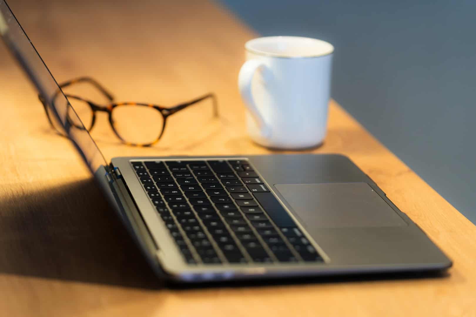 An open laptop sits on a wooden table, accompanied by a pair of eyeglasses and a white mug. Nearby, a dog gently dozes, adding warmth to this cozy setting as the sunlight bites through the window.