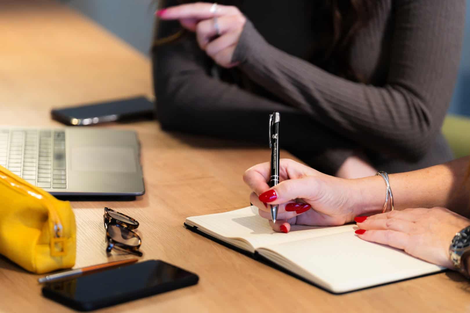 Two people sit at a table with a notebook, pen, laptop, yellow pouch, sunglasses, and a smartphone. One person is writing while the other gestures animatedly about their dog who playfully bites at the air during walks.