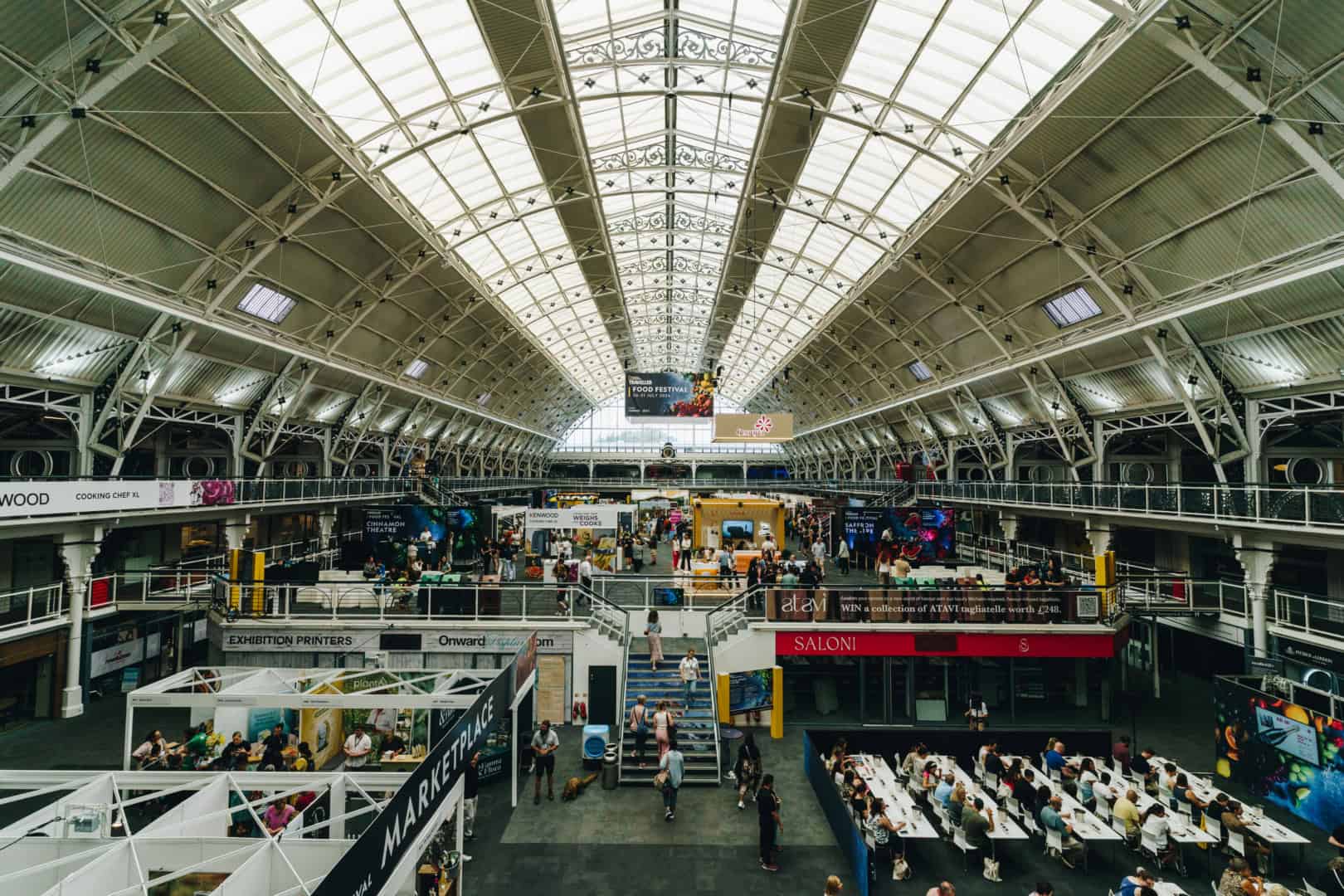 A large indoor exhibition hall with numerous stalls, people walking and sitting, and a high arched glass ceiling, offers a vibrant atmosphere reminiscent of bustling marketplaces in India. The scent of tea wafts through the air, adding to the cultural mosaic.
