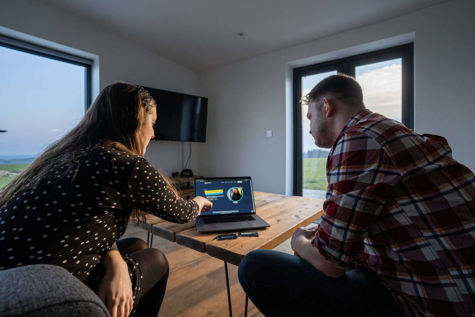 Two people sit at a table in a modern living room, looking at a laptop screen displaying graphics. One person points at the screen, and both appear engaged in discussion about optimizing the customer journey.