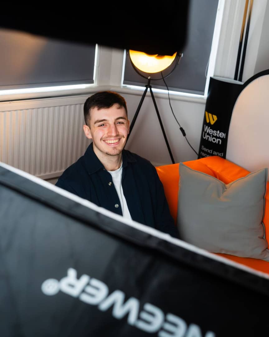 A smiling man sits on an orange couch surrounded by professional photography lighting equipment in a modern room.