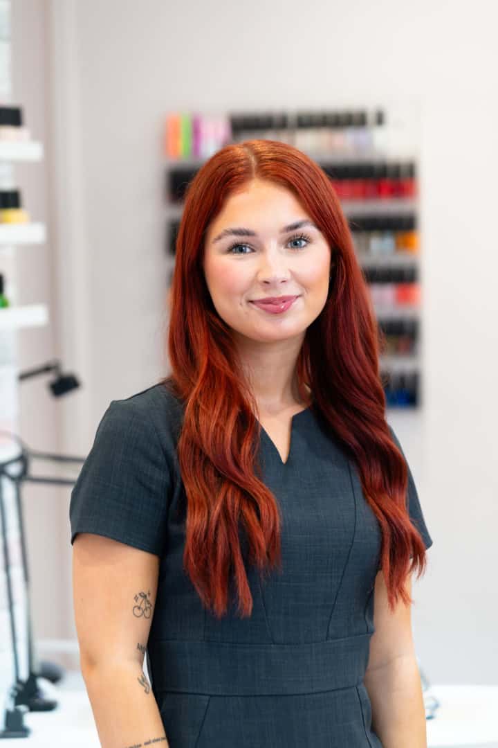 A woman with long red hair, wearing a dark short-sleeved dress, stands indoors in front of shelves filled with colorful items. The scene is illuminated by soft natural light, highlighting the vibrant details around her.