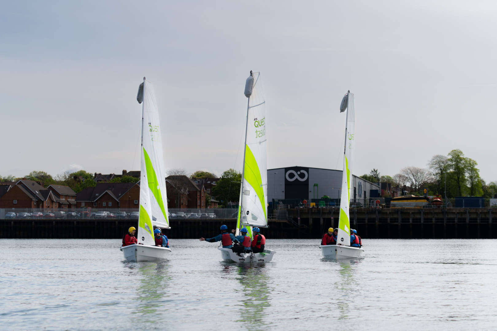 Three small sailboats with neon yellow and white sails glide across a calm body of water near a shoreline dotted with buildings, showcasing an Active Nation's spirit of sports and adventure.