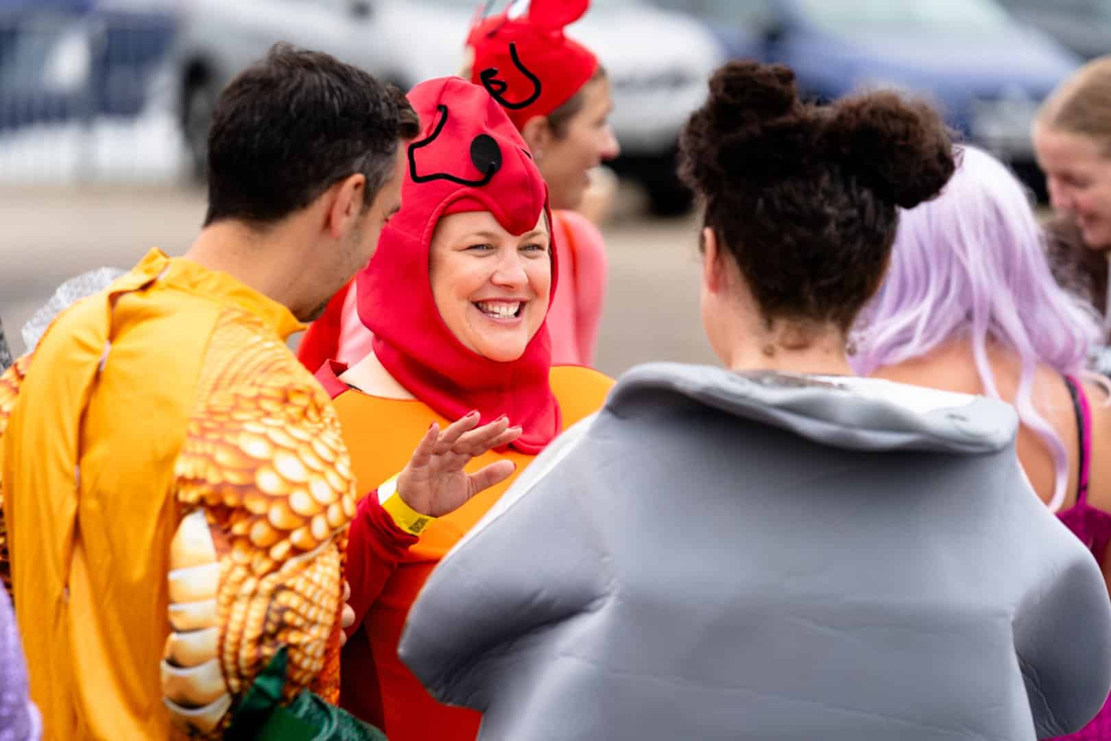 A vibrant group of people in colorful costumes, featuring a lobster and a fish, chat animatedly outdoors as excitement buzzes for the upcoming Dragon Boat Race.