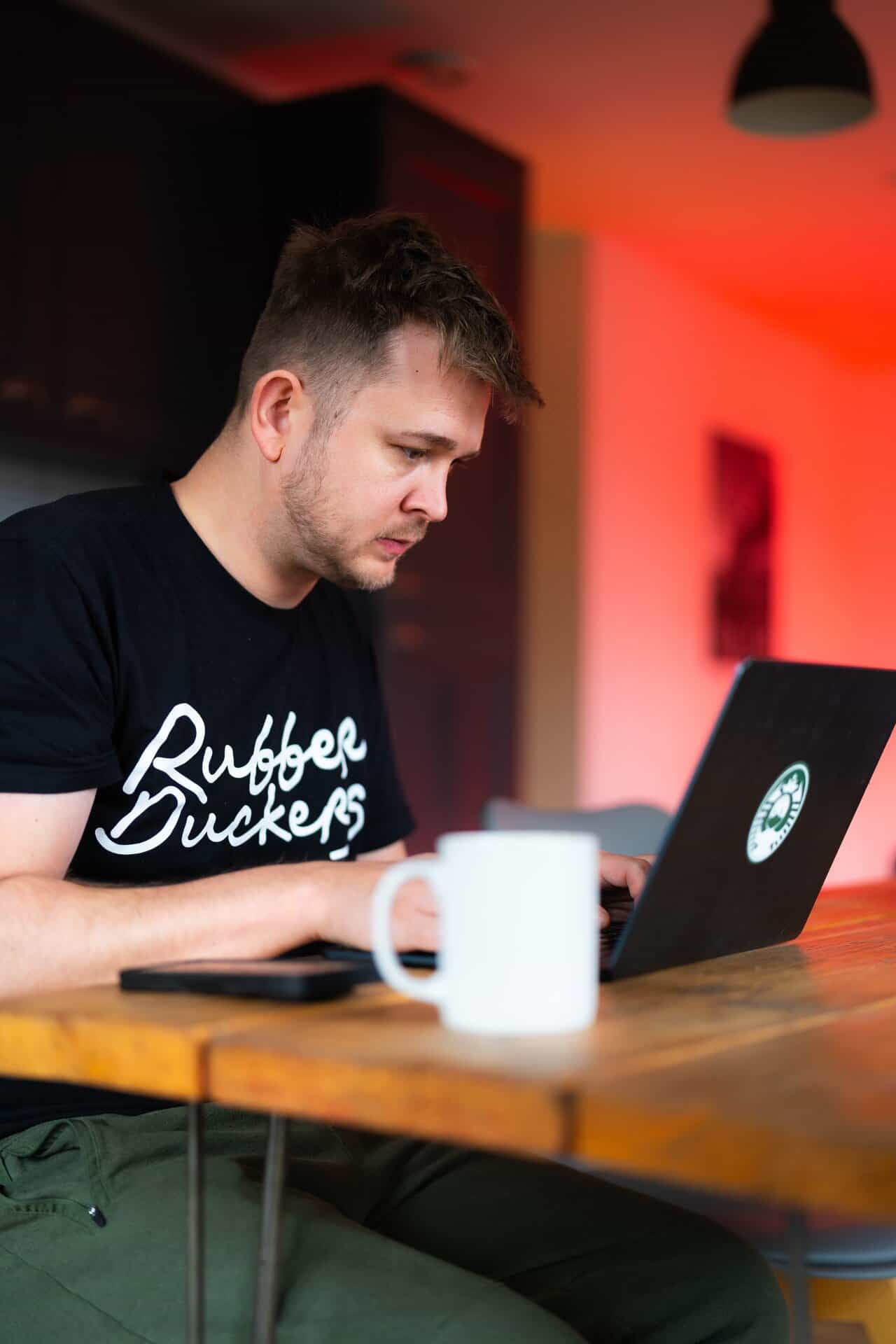 A person in a black shirt sits at a wooden table, focused on a laptop. A white mug is in the foreground. The background is softly lit in red tones. | Rubber Duckers