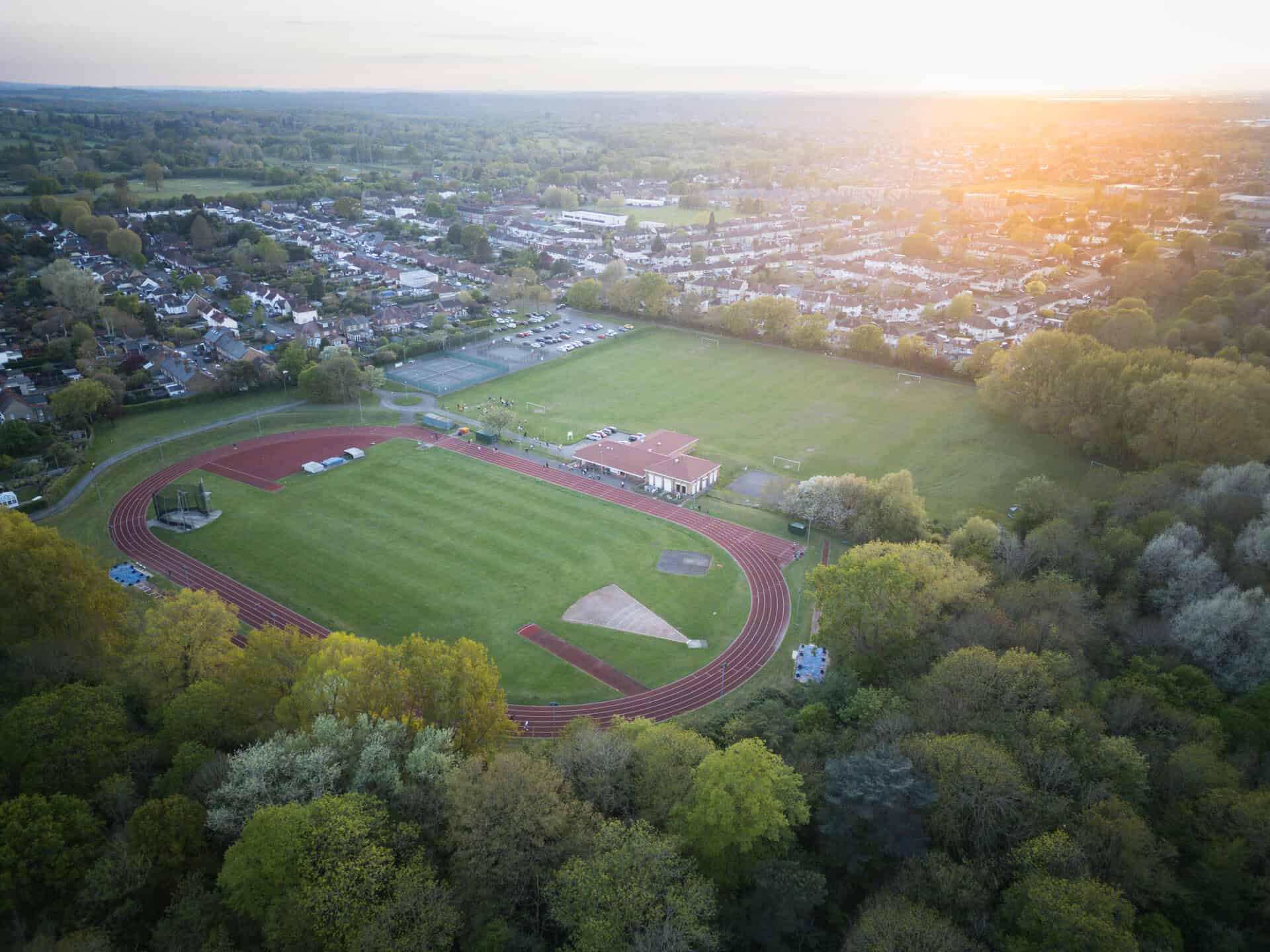 Aerial view of the Epsom & Ewell Harriers athletics club’s outdoor track and sports field surrounded by trees, with a parking lot and buildings in the background at sunset. | Rubber Duckers