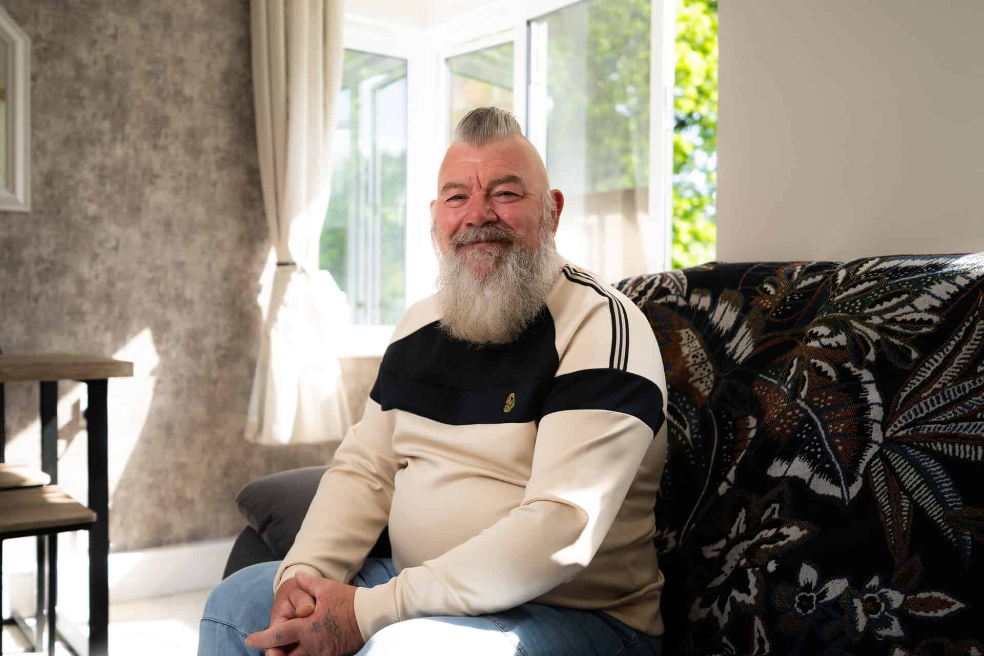 A man with a gray beard and mohawk sits on a sofa in a sunlit living room, smiling at the camera. | Rubber Duckers