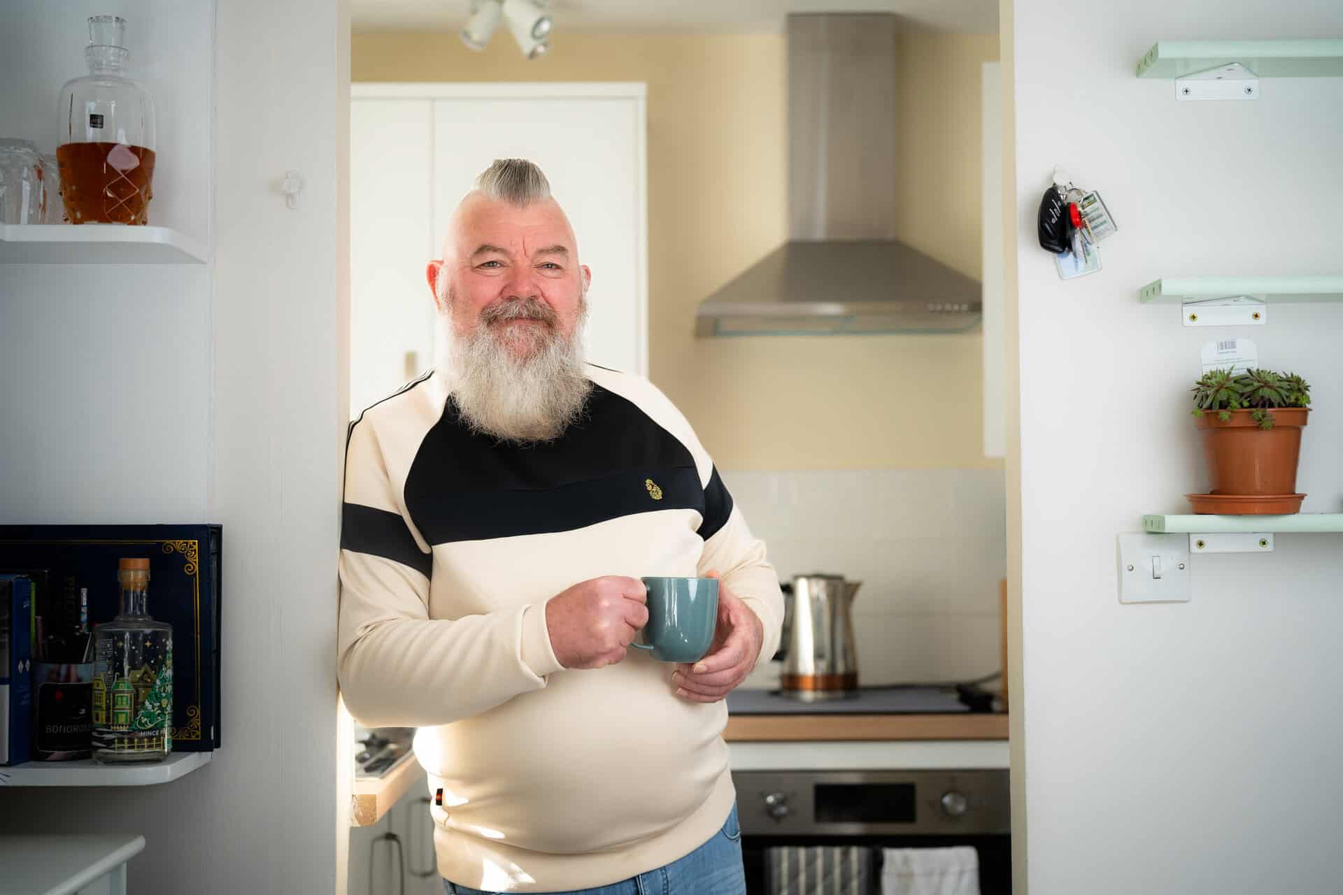 An older man with a white beard stands in a kitchen holding a mug, smiling at the camera. Shelves, a kettle, and a potted plant are visible in the background. | Rubber Duckers